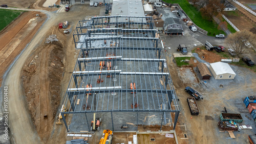 Workers are busy at a construction site erecting a large metal building. Equipment is scattered around the area, and vehicles are parked nearby. The day is clear with some clouds.