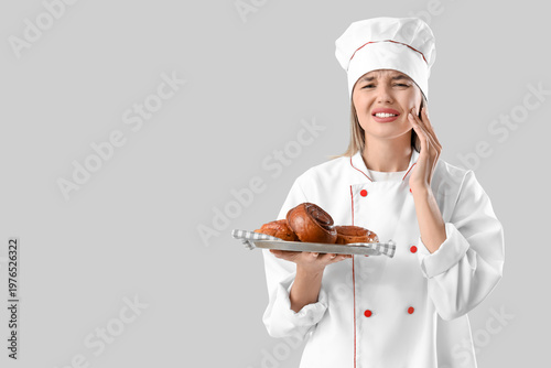 Young female chef with fresh pastry suffering from tooth ache on grey background