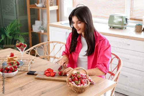 Beautiful young woman cutting fresh strawberries on table at home