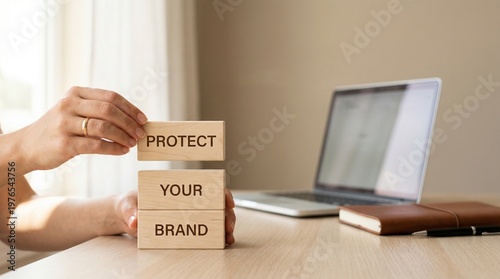 Hands arrange wooden blocks reading Protect Your Brand beside a laptop on a desk, illustrating intellectual property defense, corporate identity security, and office work setting.