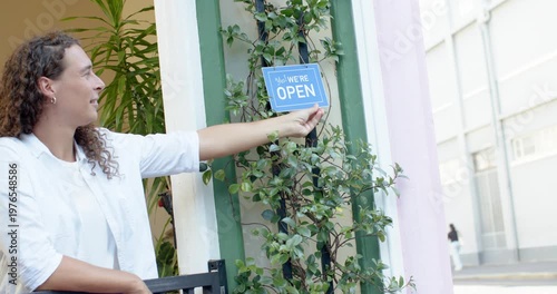Opening non-binary owner white shirt hanging blue We're OPEN sign, leaning on gate for customers