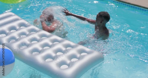 Splashing dad in green trunks and red-trunked son pushing silver float and ball yard pool playfully