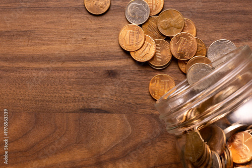 Fotografía Top-Down View of Coins Spilling from Glass Jar onto Wooden Surface