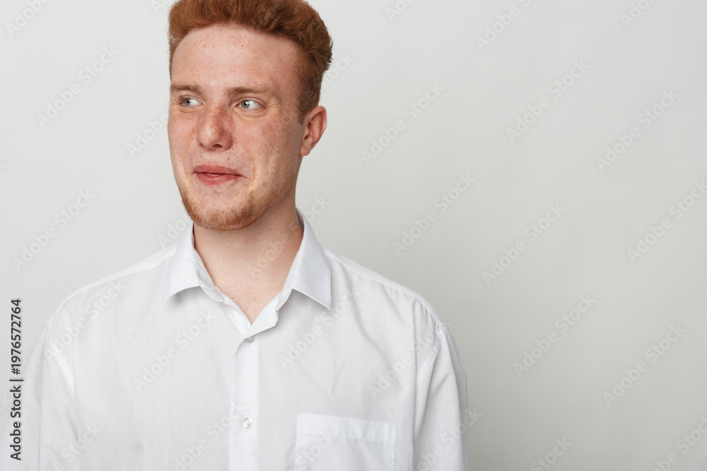 Obraz premium Young man with red hair and freckles wearing white shirt looks sideways with thoughtful expression on plain light background. Concept of casual style, contemplation, and natural appearance.