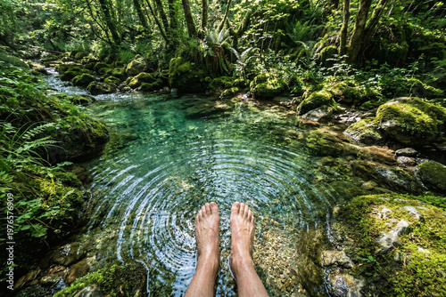 Point of view of bare feet dipping into a crystal clear stream creating ripples in a lush green mossy forest
