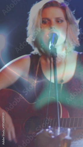 Vertical video: Hand with glass revealing musician strumming guitar, performing at mic under flares