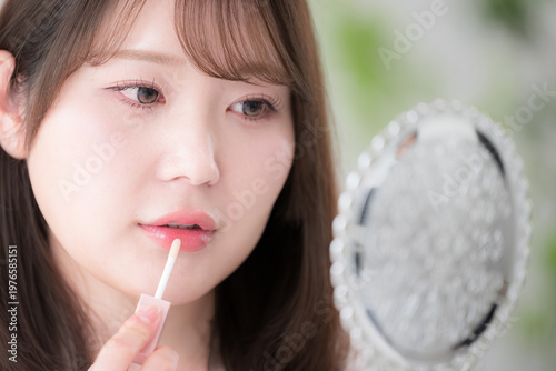 手鏡を見ながらリップを塗る女性のナチュラルメイクと美容ケア　リップグロスや唇のケアなどのイメージクローズアップ