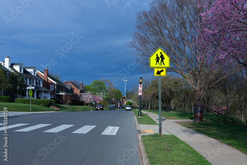 Residential neighborhood in Frederick, Maryland, with yellow school crossing sign and pink spring blossoms under a dark storm sky on S Jefferson Street.