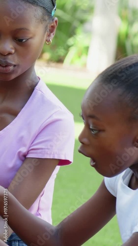 Vertical video: Giving pink-tee handing black pot to white-tee girl in yard sharing yellow blooms