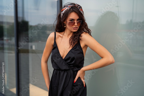 Beautiful fashion girl in black summer dress with sunglasses and hair bandana posing in front of store window, looking at camera 
