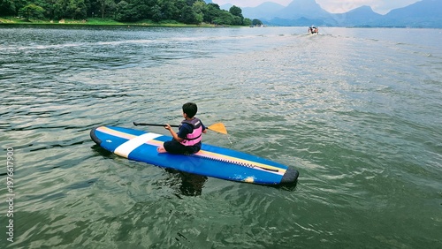 An East Asian boy is sitting on stand up paddle board at a beautiful lake under blue sky on with amazing hills and mountain in background.