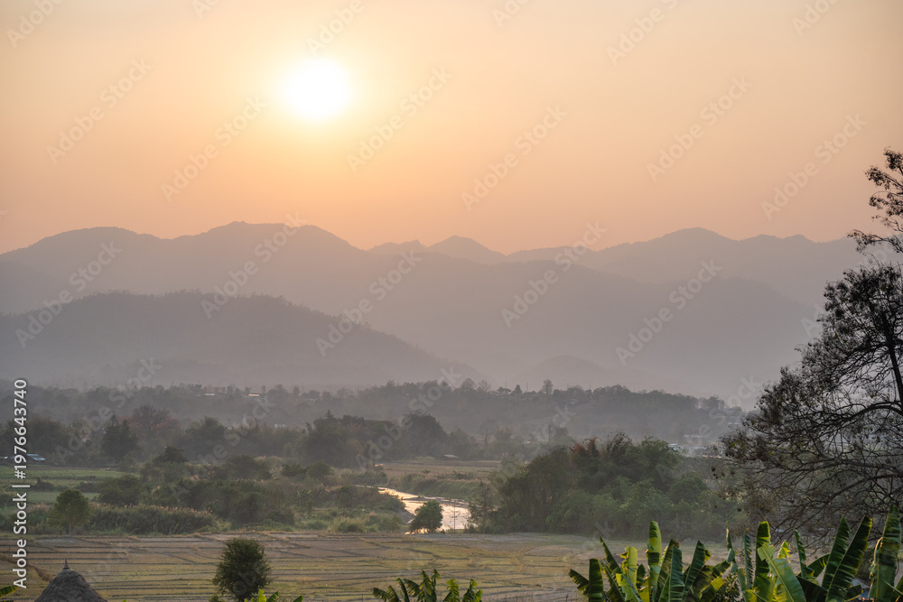 Fototapeta premium Exotic rural plantations and rice fields landscape of Pai city and river in Northern Thailand with fog from forest fires covering hills and meadows at sunset, sun in smog shining at countryside.