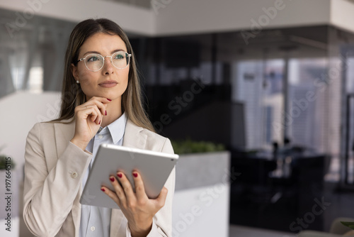 Canvas Print Thoughtful professional woman stands in modern office, holding digital tablet, staring away with pensive expression, looks in deep concentration or decision-making