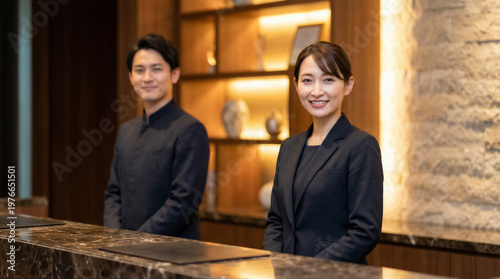 Smiling hotel staff members, a man and a woman, standing behind the reception desk in a modern hotel lobby