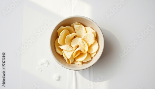 A simple bowl of potato chips rests on a white tablecloth in warm natural sunlight, conveying a casual snack moment, texture, and minimalist home dining atmosphere ideal for food and lifestyle project