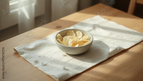 A simple bowl of potato chips rests on a white tablecloth in warm natural sunlight, conveying a casual snack moment, texture, and minimalist home dining atmosphere ideal for food and lifestyle project