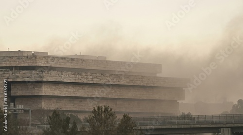 Layered brutalist structure half erased by dusty atmospheric veil beyond a low bridge, presenting institutional mass through polluted light, soft contrast, and urban environmental strain