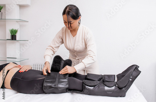 Therapist Operating Pressotherapy Machine While Female Client Lies with Full Leg Air Compression Boots on Massage Table — Professional Pneumatic Lymphatic Drainage Treatment for Legs, Anti-Cellulite 