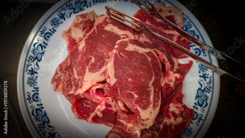 Raw marbled beef slices on vintage ceramic plate with serving tongs, fresh uncooked red meat preparation, rustic kitchen food photography, butcher cut steak ready for cooking, traditional home cooking
