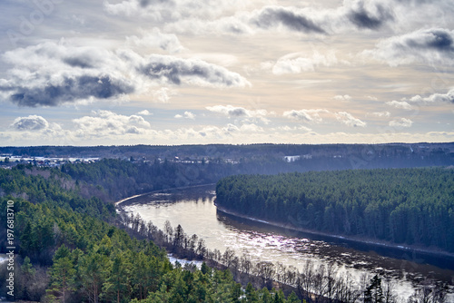 A panoramic aerial landscape view capturing a wide river winding through dense evergreen forests in Birštonas, Lithuania. The river, partially covered with ice and snow, curves gracefully between
