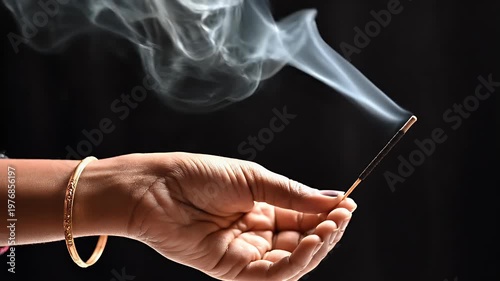 A hand delicately holds a burning incense stick against a dark background, releasing fragrant smoke into the air.