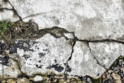 top view of heavily damaged light concrete pavement with deep cracks and black tar spots. The texture features organic debris, green grass, and stone aggregate underneath, representing infrastructure 