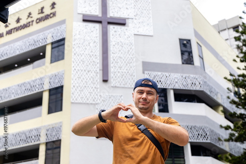 Man in orange t-shirt making heart shape with hands in front of a modern Methodist church building with a large cross in Singapore. Urban travel and religious faith context.