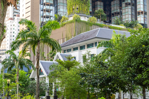 Modern skyscraper featuring vertical greenery and hanging gardens behind a traditional white building in Singapore urban landscape during daytime. Sustainable architecture and biophilic design.
