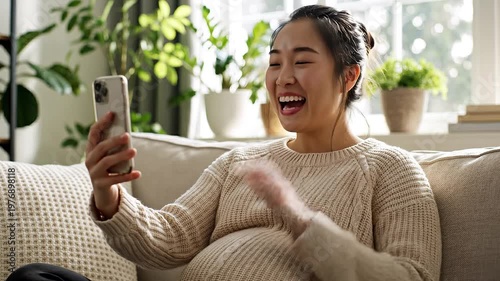 Happy pregnant woman relaxing at home while using smartphone and enjoying video call in cozy living room space