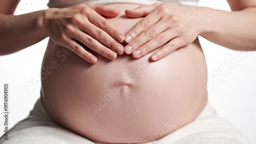 Close-up of a pregnant woman gently caressing her bare belly while sitting against a white background