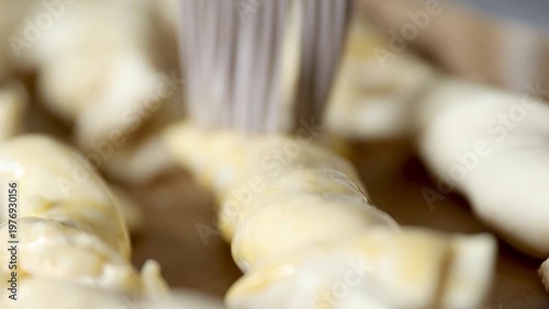 Baker applying egg wash with a brush on uncooked pastry dough pieces arranged on a baking tray in a well-lit kitchen setting