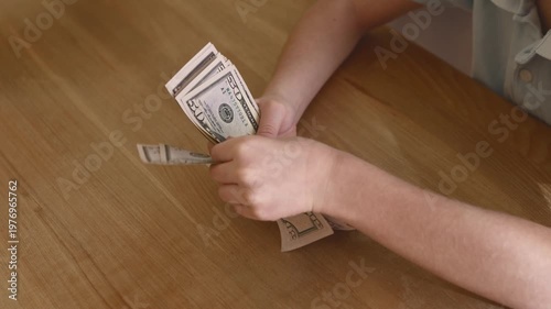 Child counting thick stack of US dollar bills on wooden desk, kid sorting cash banknotes, teaching kids about budgeting and saving money, indoor close-up hands with money