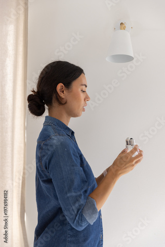 LED lightbulb preparing to be installed into empty lamp socket on white wall near beige curtain