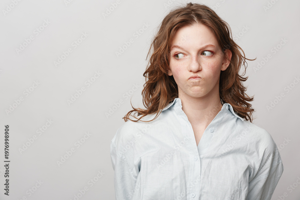 Obraz premium Young woman with curly hair in white shirt looks sideways with skeptical expression on light background. Concept of doubt, suspicion, and uncertain emotions.