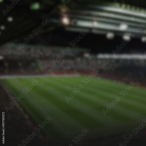 An abstract, out-of-focus view of a professional football stadium during a night match. The image shows a lush green pitch under intense stadium lights with a blurred audience in the stands. This boke