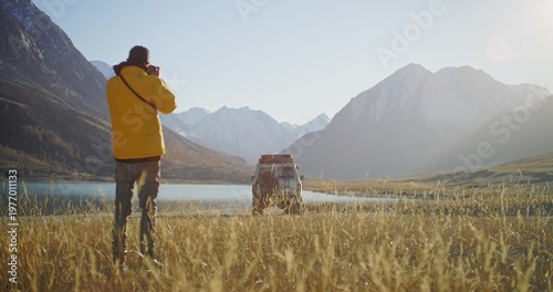 Photographer in a yellow jacket taking pictures near an off road SUV parked by a mountain lake in a remote alpine valley. Golden grass, snowy peaks, and bright sunlight create a cinematic outdoor