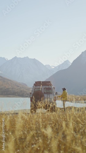 Traveler in a yellow jacket getting into an off road SUV parked by a mountain lake in a remote alpine valley. Golden grass, snowy peaks, and bright natural light create a scenic outdoor travel scene