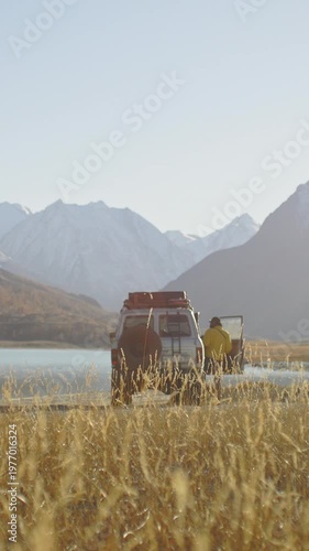 Traveler in a yellow jacket carrying a camera and walking toward an off road SUV parked by a mountain lake in a remote alpine valley. Golden grass, snowy peaks, and bright sunlight create a scenic