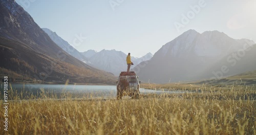 Photographer in a yellow jacket standing on the roof of an off road SUV and shooting mountain scenery by a calm lake in a remote alpine valley. Golden grass, snowy peaks, and bright sunlight create a