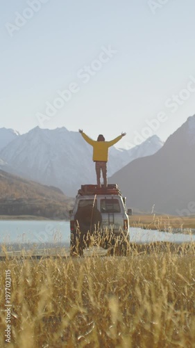Traveler in a yellow jacket standing on the roof of an off road SUV with raised arms near a mountain lake in a remote alpine valley. Snowy peaks, golden grass, and bright sunlight create a powerful