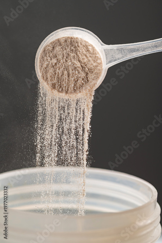 close-up shot of a woman's hand using a plastic scoop to pour whey protein powder into a transparent shaker bottle. A silver supplement container and a black lid are on a white table against a grey ba