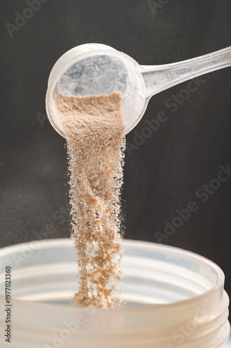close-up shot of a woman's hand using a plastic scoop to pour whey protein powder into a transparent shaker bottle. A silver supplement container and a black lid are on a white table against a grey ba