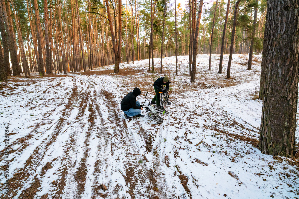 Fototapeta premium Two Young Men Preparing and Repairing BMX Bikes on a Snowy Path in a Pine Forest During Winter
