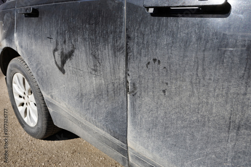 Dirty black car covered with dust and dried mud outdoors. Side mirror and door with heavy contamination, neglected vehicle surface, lack of maintenance and cleaning
