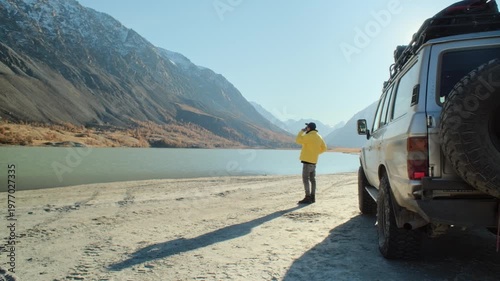 Male traveler in a yellow jacket drinking coffee from a mug beside an off road SUV parked near a mountain lake. Snowy slopes, sandy shore, and bright daylight create a commercial outdoor travel scene