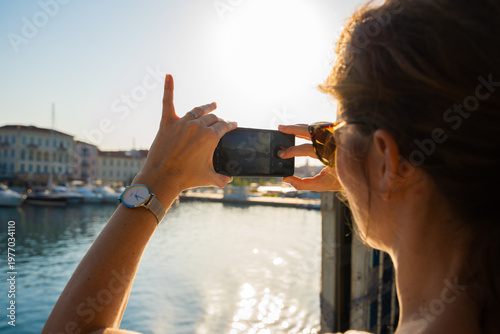 Venice, Italy - 29 March 2026: View of a woman capturing a photo of the canal's glimmering waters and historic buildings under the bright, warm sunlight.