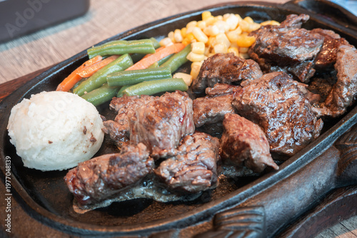 Sizzling diced beef steak with vegetables on black iron plate.