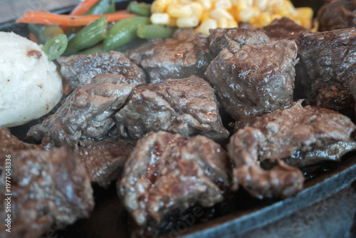 Sizzling diced beef steak with vegetables on black iron plate.