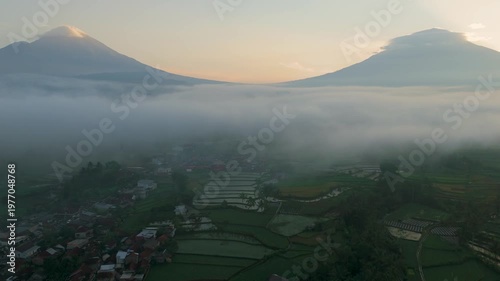 A cinematic sunrise scene over Mount Sindoro and Mount Sumbing in Central Java, Indonesia, with thick morning mist covering the valley and rice fields below.