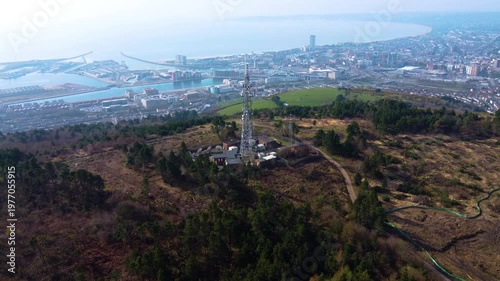 Aerial View of TV Radio 5G Transmitter Tower High Above Swansea City with Marina in Distance. Communication Infrastructure for Smart Phones and Connectivity.